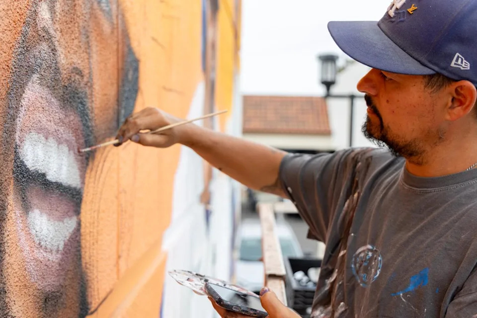 A man in a baseball cap and faded T-shirt holds a palette in one hand and a brush in the other while painting part of a mural