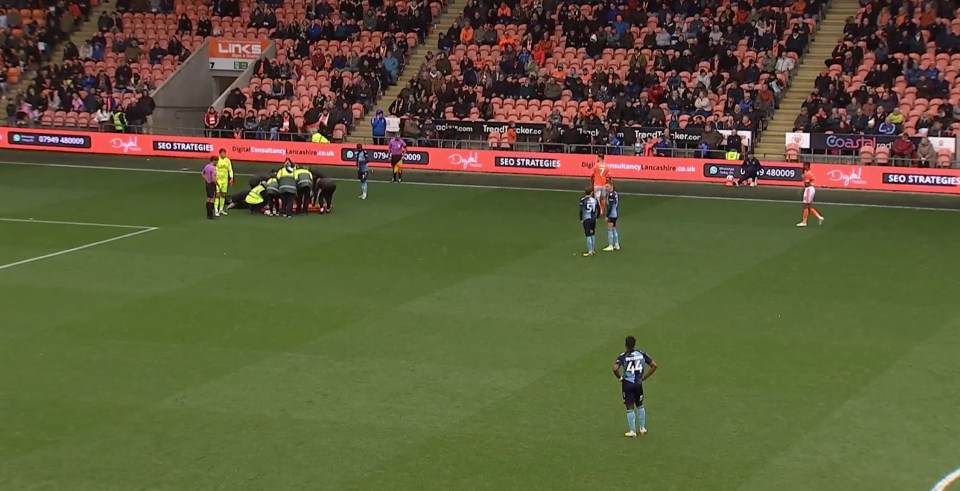 A soccer game with the scoreboard showing BPL 1-0 WYC at 89:58, as players attend to an injured player on the field.