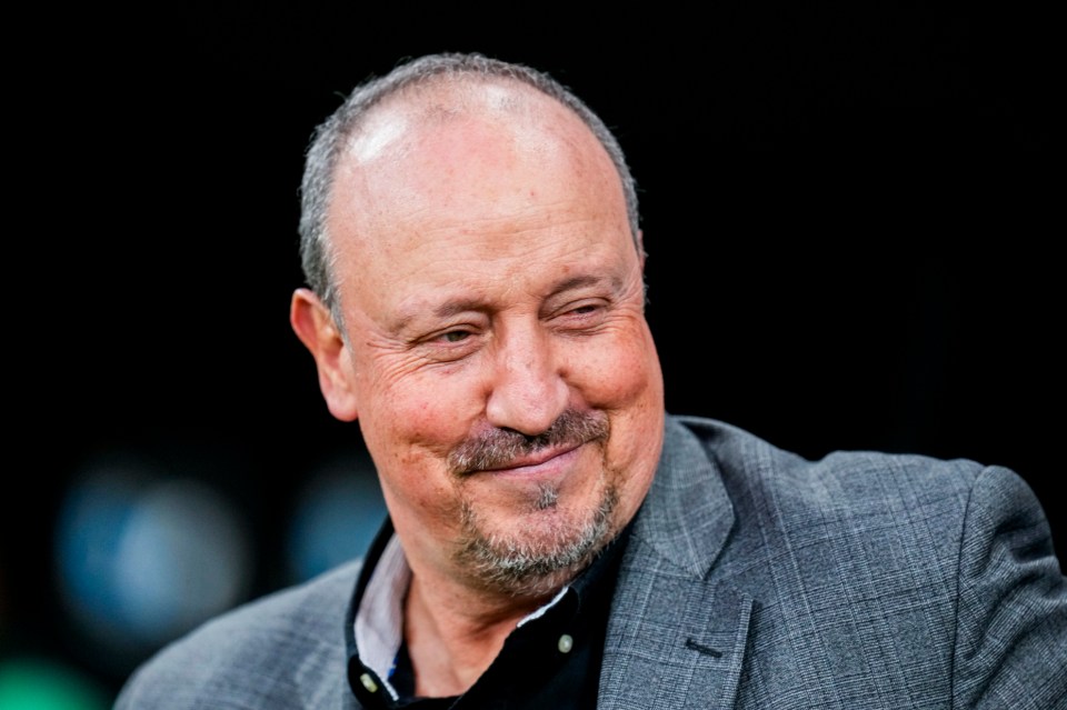 A smiling man with a graying beard and receding hairline, wearing a gray plaid suit jacket over a black shirt.