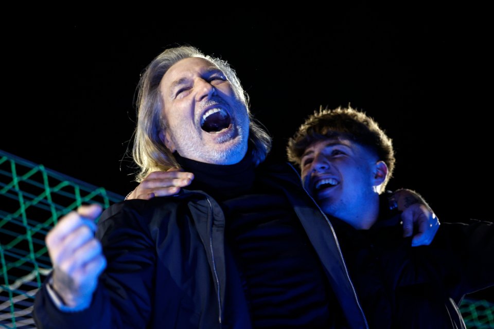 A man with long blonde hair and a younger man with curly brown hair celebrate joyously, their mouths open in excitement, under blue light at a football match.
