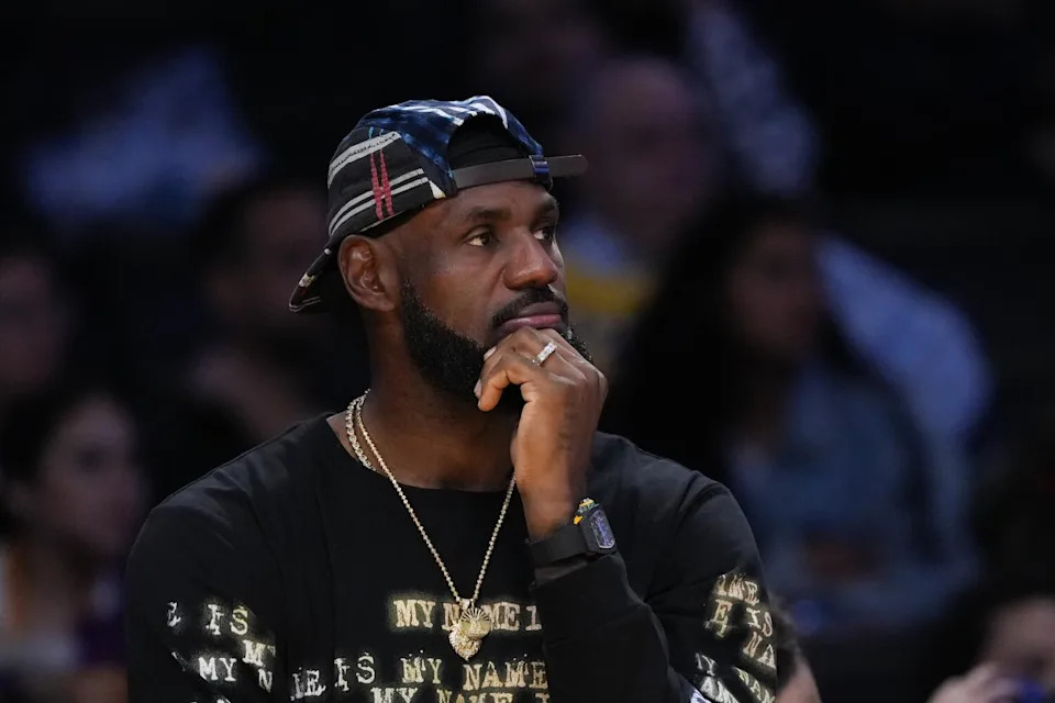 Lebron James wears a backwards cap and holds his left hand to his chin as sits on the Lakers bench.