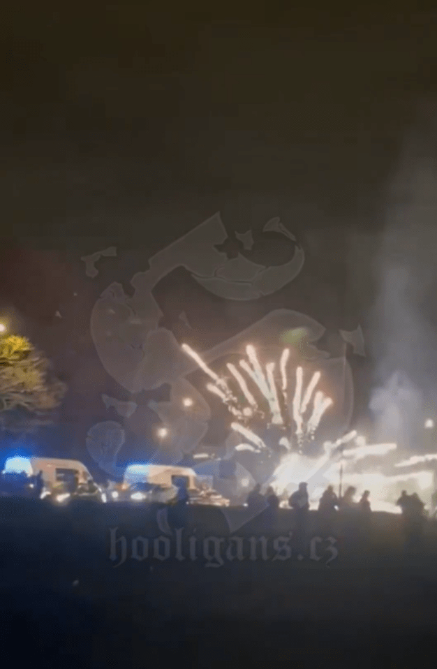 People gather on a street with flares and fireworks at night, with white vans and emergency lights visible in the background.