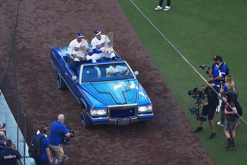 The Los Angeles Dodgers' Freddie Freeman and Miguel Rojas carry the 2024 and 2025 World Series trophies in a car driven by actor Will Farrell before the Opening Day game against the Arizona Diamondbacks at Dodger Stadium.