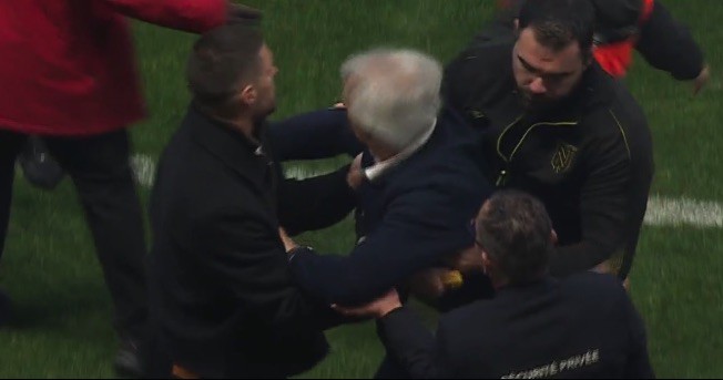 Several men in jackets physically restraining a white-haired man on a grassy field.
